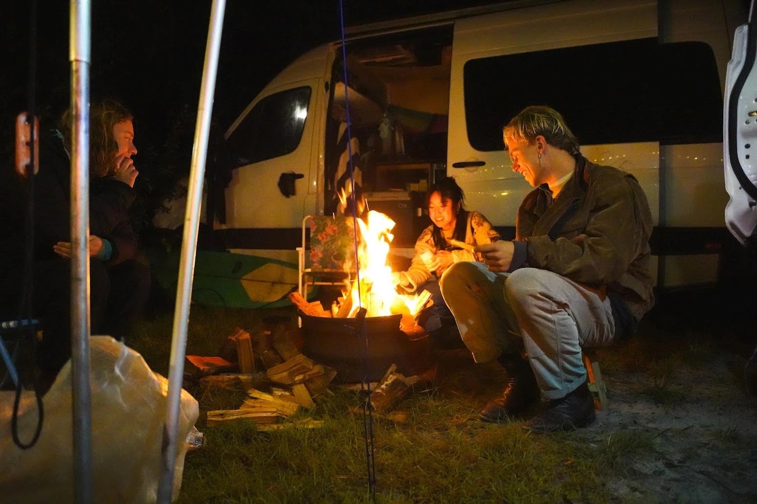 Three friends around a fire pit at night beside an open campervan