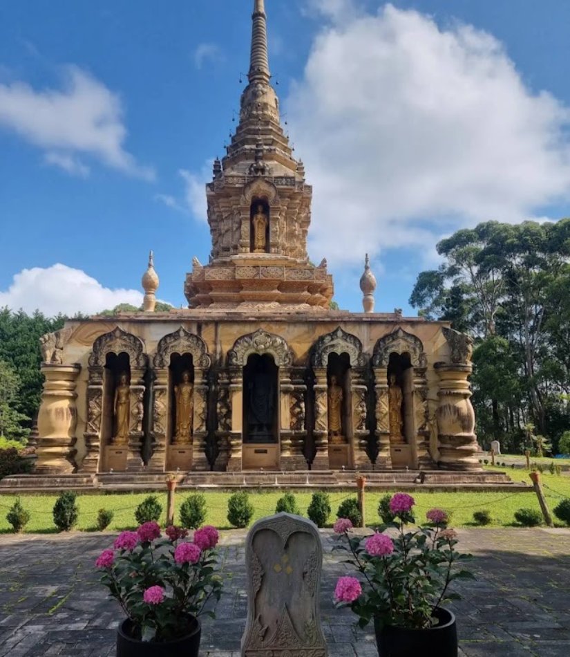 An ornate golden Thai stupa with standing Buddha statues in its alcoves, under a blue sky with white clouds, pink flowers in the foreground