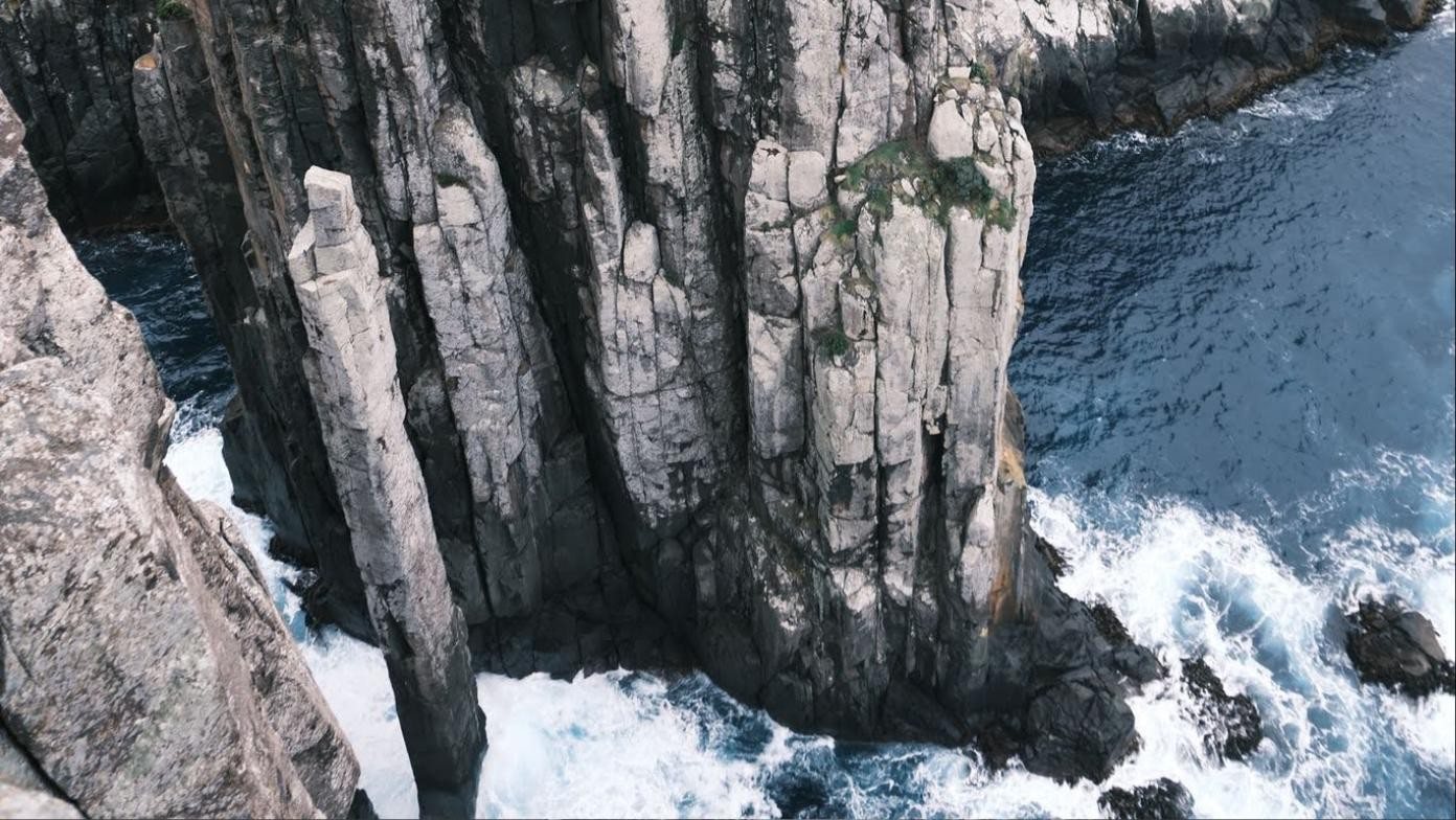Two sea stacks of pale columnar rock, one slender, one broad, standing in white surf below a cliff