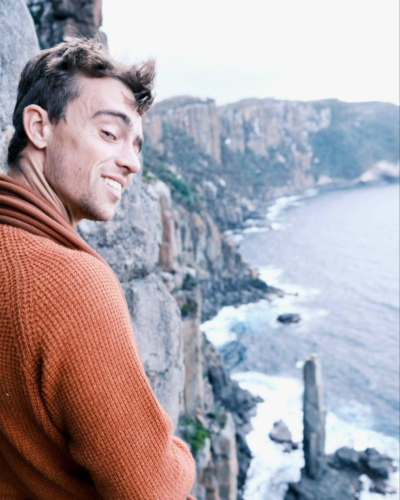 Sam in an orange jumper on a cliff edge, a coiled rope in his hand, sea stacks and surf far below