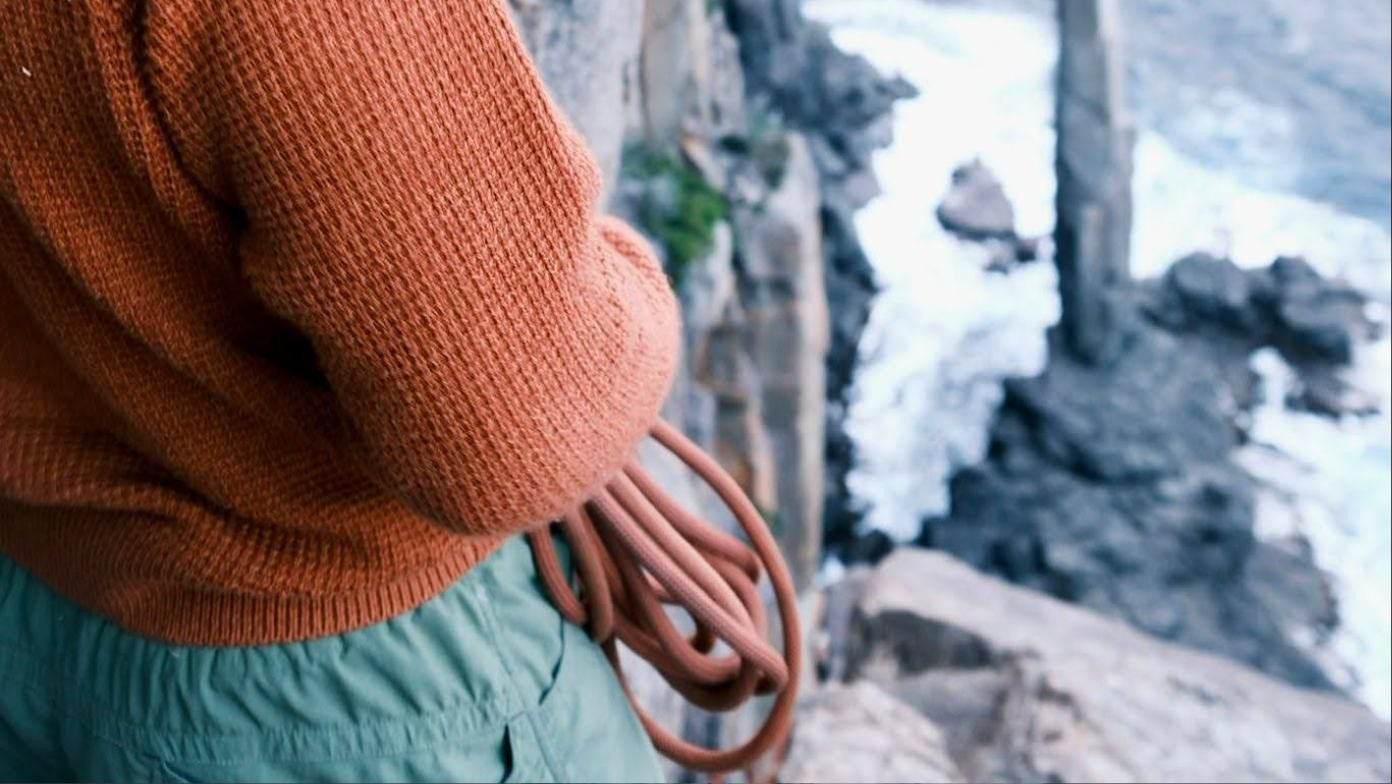 A coiled climbing rope held at the hip above a drop to rocks and white water