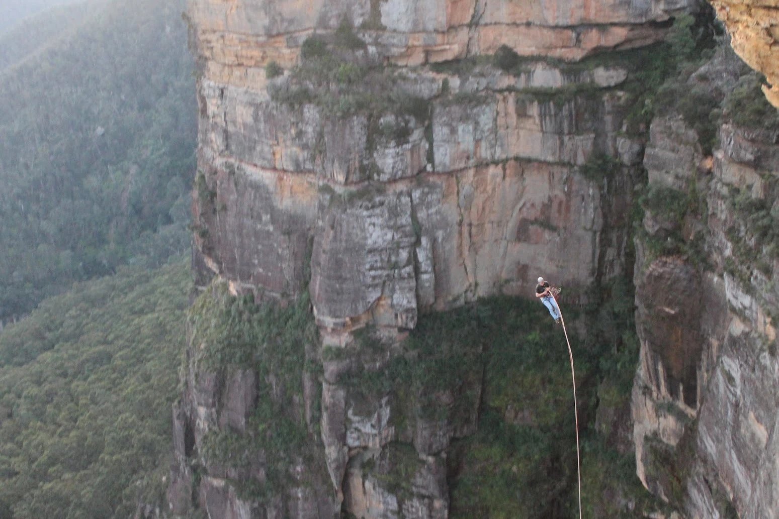 A small figure hanging on a single long rope in front of a towering sandstone cliff above a forested valley