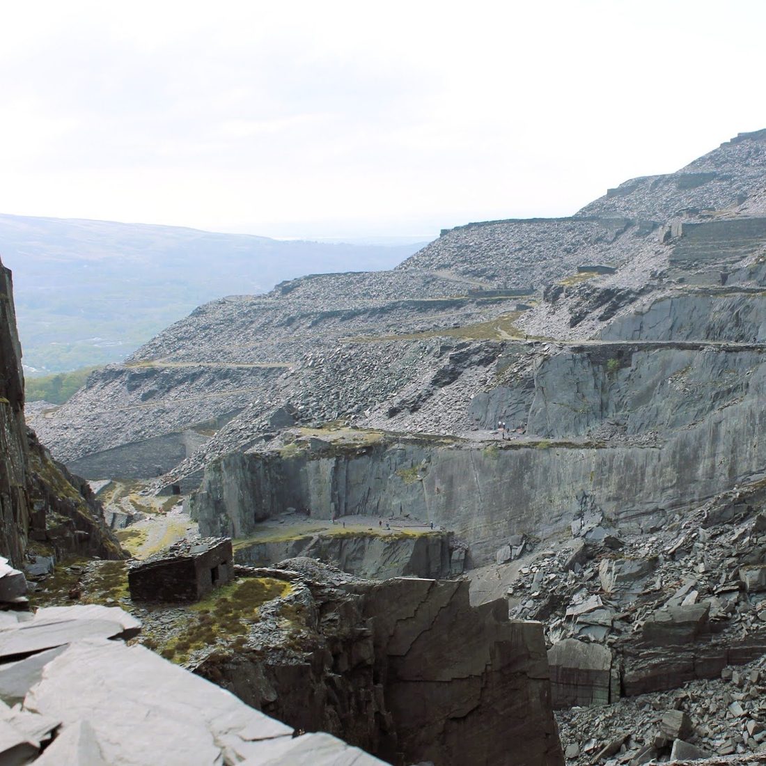 Terraced slate quarry walls and spoil heaps in grey light, with hills beyond