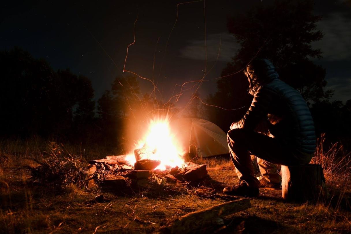 A figure in a hooded jacket sitting alone on a log beside a campfire at night, sparks rising, a tent behind