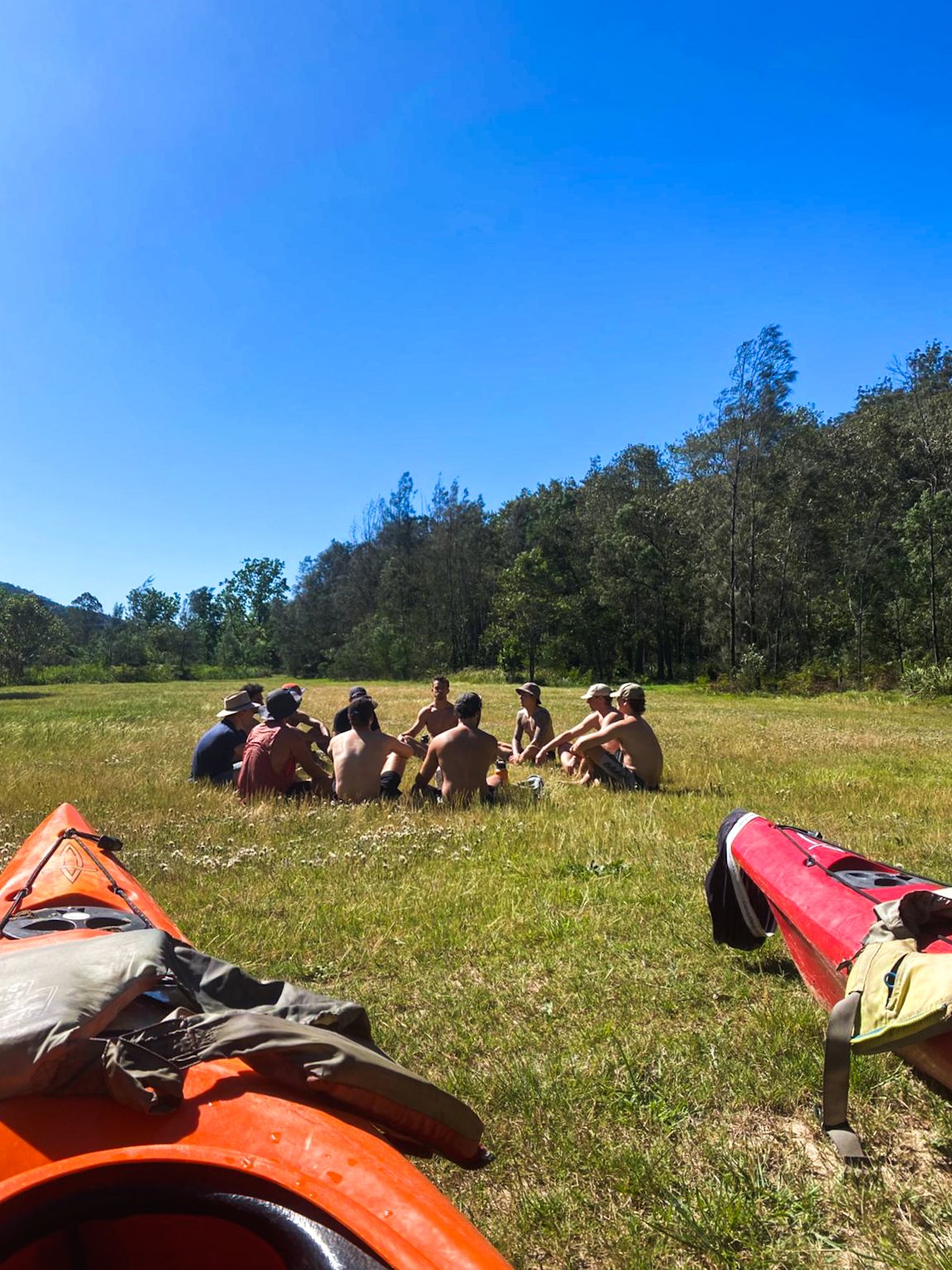 A group of men sitting in a circle in a wide grassy field, two kayaks in the foreground, forest behind