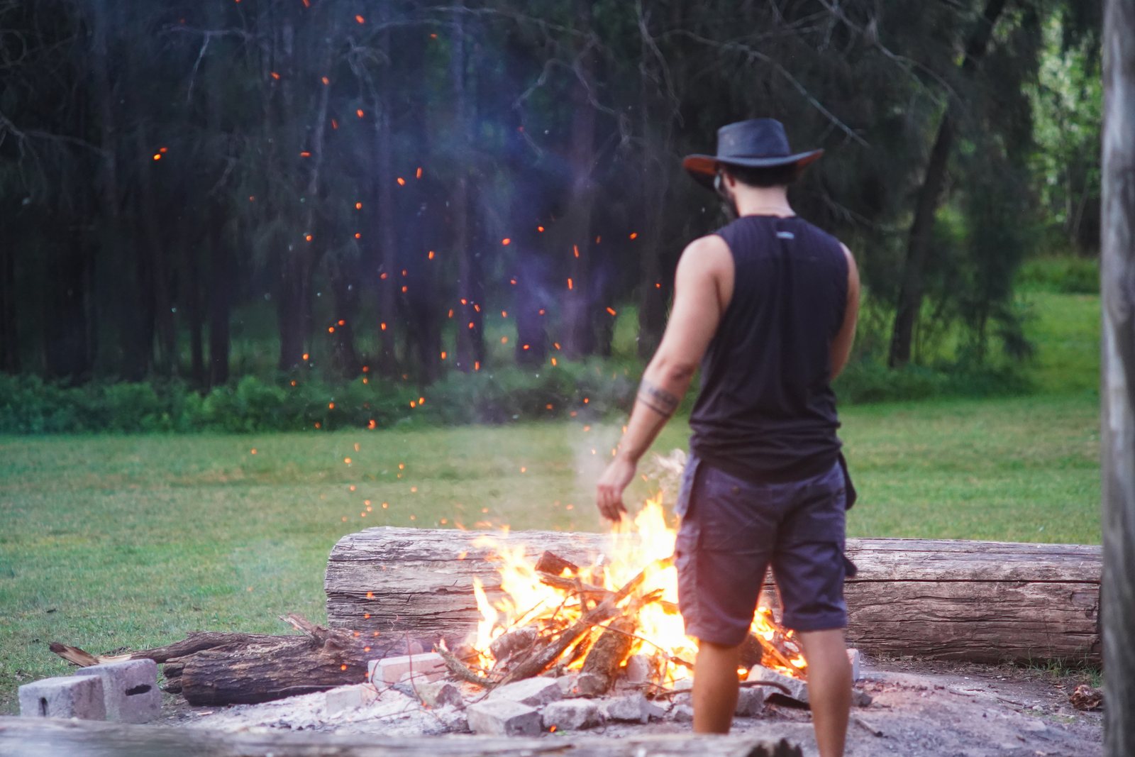 A man seen from behind, in a hat and singlet, stoking a large campfire as sparks rise into the trees