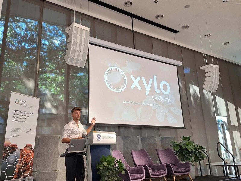 Sam presenting at a lectern in front of a large screen reading Xylo Systems, Species Occurrence Modelling, with a DARE Centre banner beside the stage