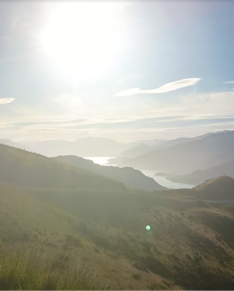 Hazy green hills falling away towards a distant lake, with the sun low and bright above them