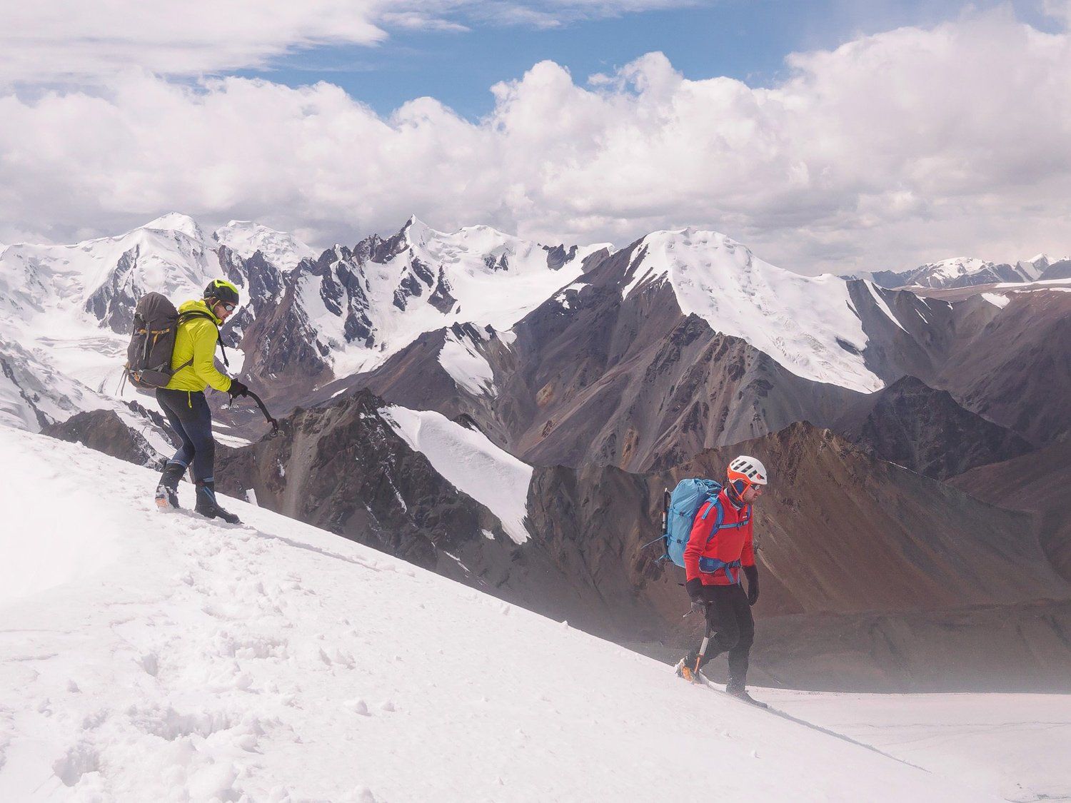 A climber in a red jacket on a steep snow slope below rock and cloud