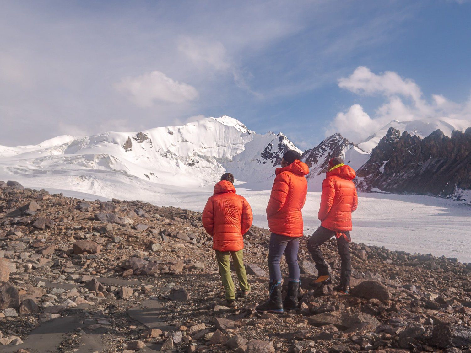 Three mountaineers in orange jackets looking out across a glacier to snow peaks